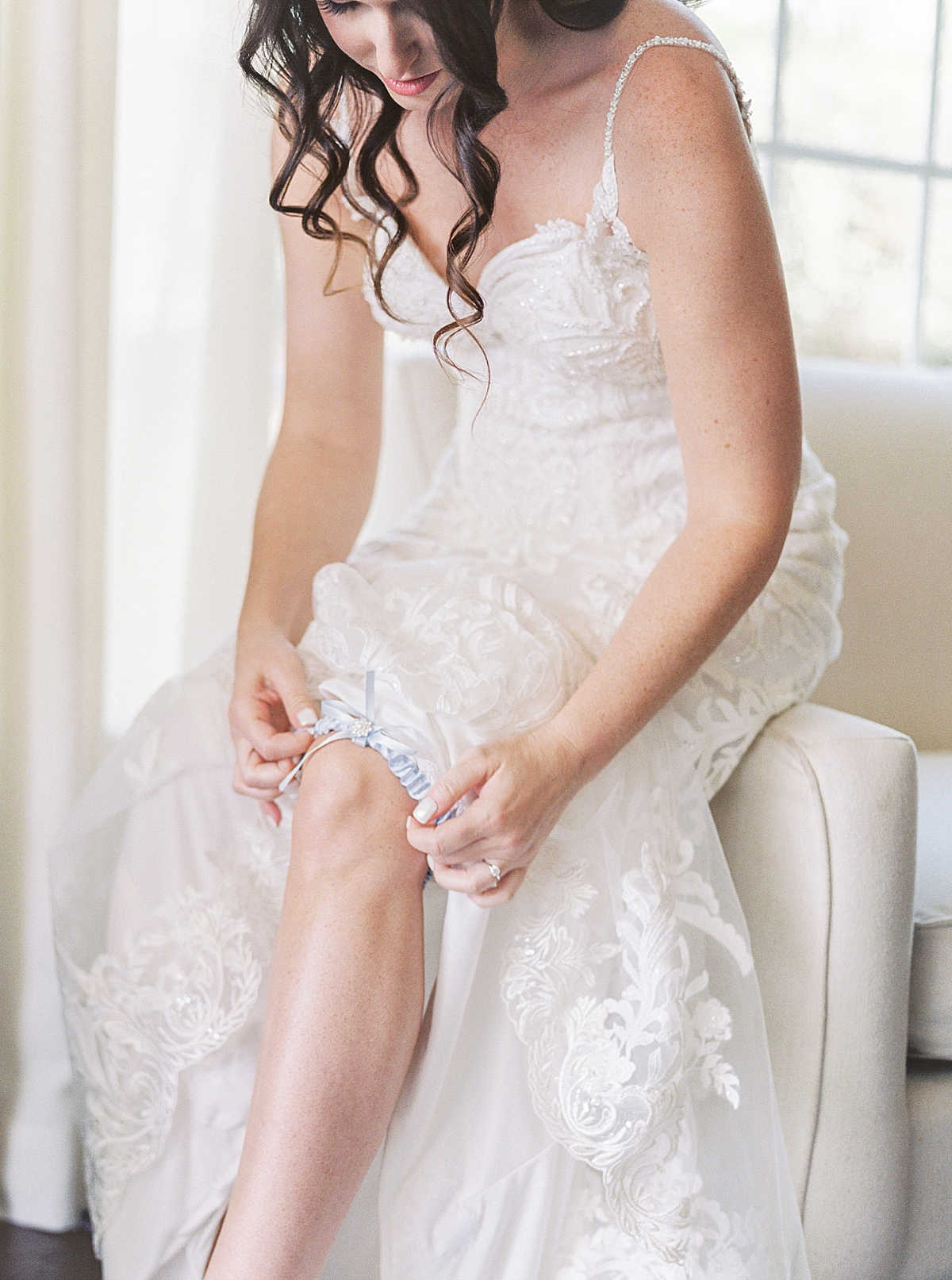 Bride in sparkly lace wedding dress with long lace train putting on blue garter, something blue wedding