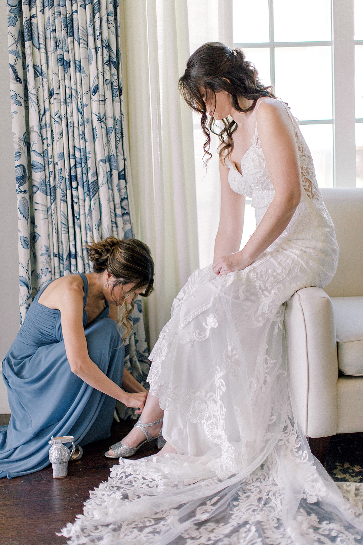 Bride in lacy wedding dress getting ready putting on shoes, bridesmaid in dusty blue bridesmaid dress