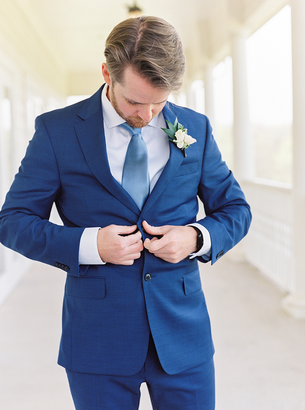 Groom getting ready in blue suit with light blue tie, white boutonniere on porch of milestone mansion