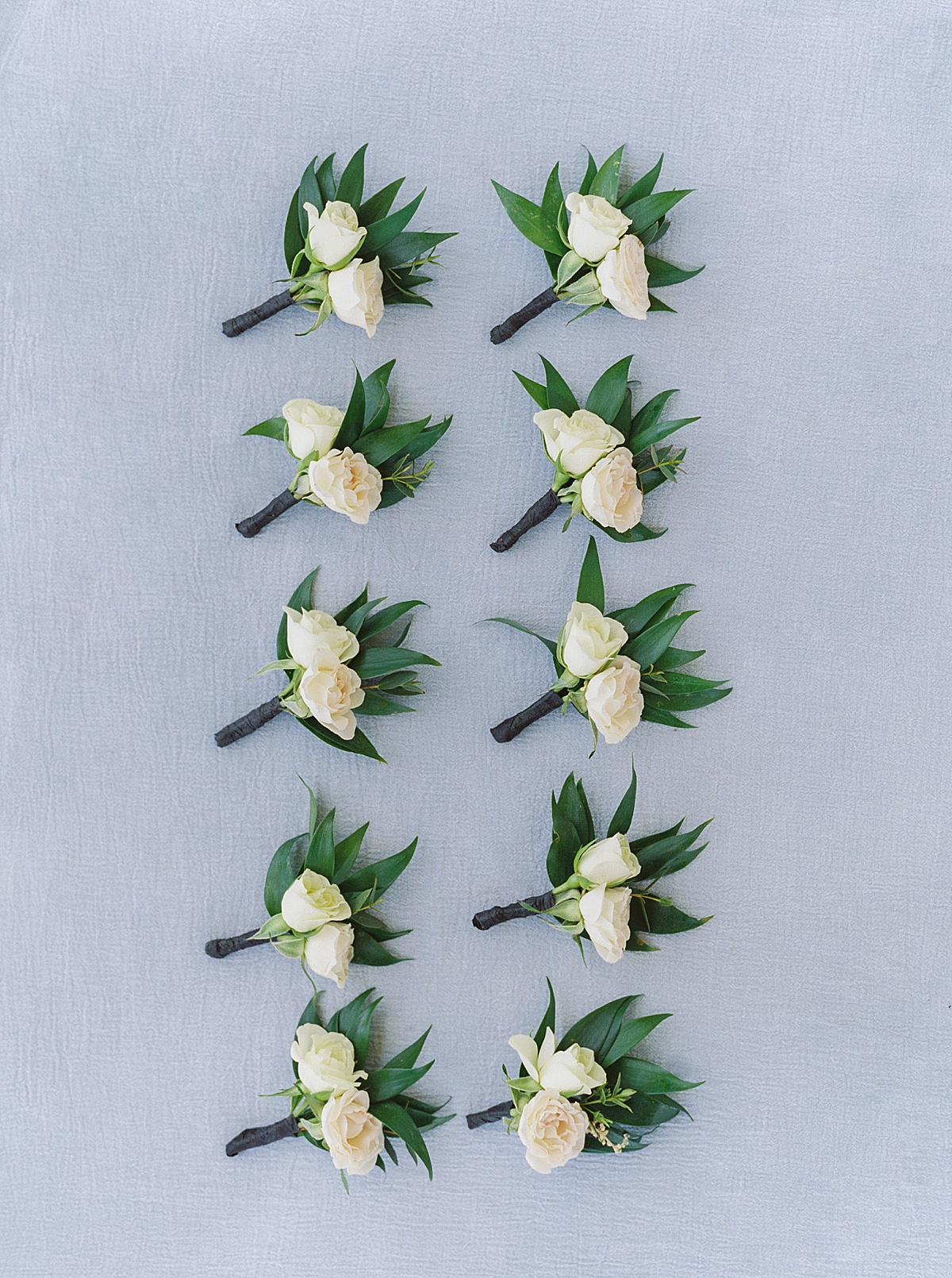 groom and groomsmen boutonnieres on light blue background, white wedding flowers, white rose boutonniere with greenery