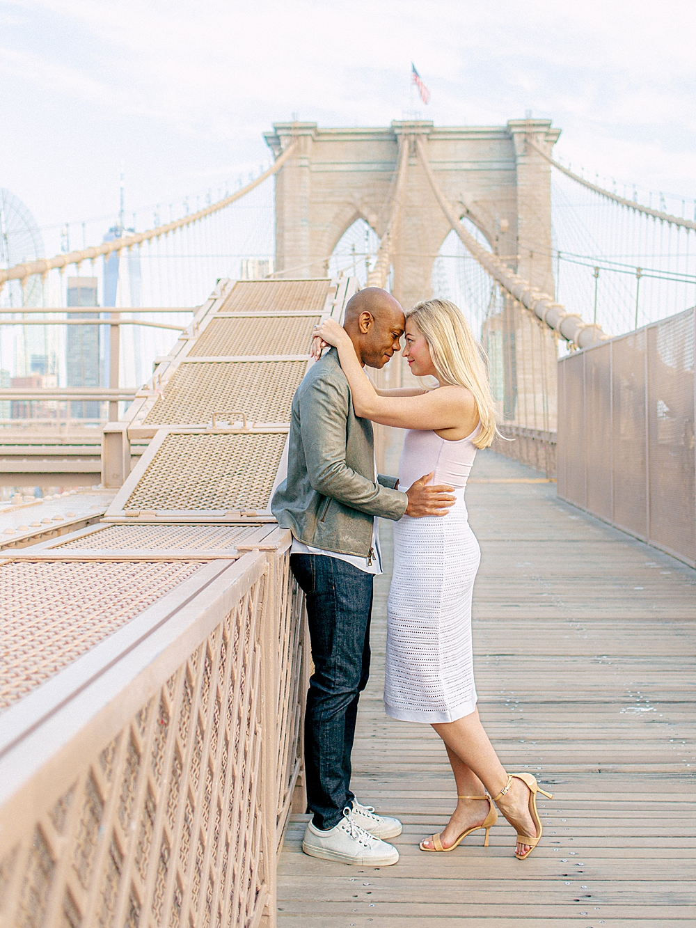 Engaged couple taking engagement photos on the Brooklyn Bridge wearing a white dress and gray blazer with a white shirt and jeans, New York City wedding and engagement photographer
