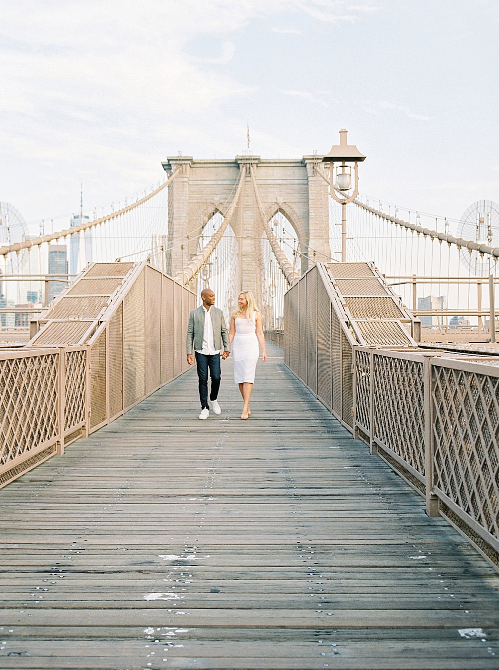 Engaged couple walking on the Brooklyn Bridge wearing a white dress and gray blazer with a white shirt and jeans, New York City wedding and engagement photographer